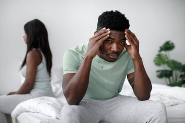 Bir adam yatağın kenarına oturmuş, stresli ve düşünceli görünüyor. Ortağı yakınlarda bir yerde. İkisi de sakin ve tarafsız bir odada ilişkilerini yansıtıyor..