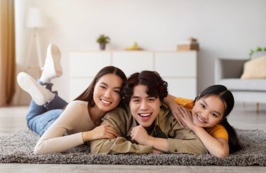 Üç arkadaş yumuşak bir halının üzerinde uzanıyor, gülümsüyor ve birbirlerini mutlu bir şekilde kucaklıyorlar. Parlak oturma odası doğal ışık ile sıcak bir atmosfere sahiptir..