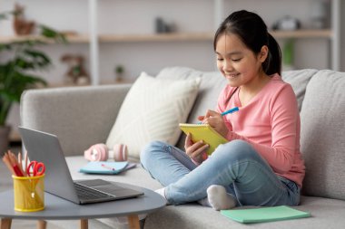 İnternetten öğrenme seansına katılan bir kız kanepeye oturur. Okul malzemeleri ve dizüstü bilgisayarla çevrili not defterine notlar yazarken gülümsüyor..