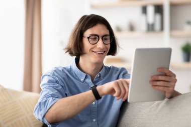 Uzun kahverengi saçlı ve gözlüklü genç bir adam rahat bir oturma odasında tabletle etkileşime girerken gülümsüyor. Doğal ışık uzayı aydınlatır ve sıcak bir atmosfer yaratır..