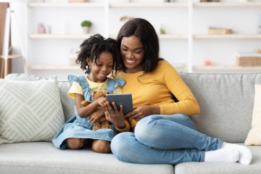 Bir anne ve kızı rahat bir kanepede oturur, gülümser ve tablete bakarlar. Kız bir oyuncak ayı tutuyor ve evlerinde neşeli bir anı paylaşıyorlar..