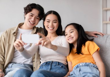 Üç aile üyesi akıllı telefondan selfie çekerken mutlu bir şekilde gülümsüyor. Havalı atmosfer, şık bir oturma odasında güçlü bir bağ olduğunu gösteriyor..