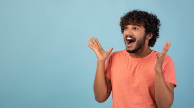 Pembe tişörtlü kıvırcık saçlı Hindu adam neşeli bir ifadeyle yan tarafa bakıyor. Mavi arkaplanı olan bir stüdyoda elleriyle jest yapıyor, heyecan gösteriyor..