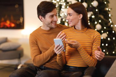 Genç bir çift rahat bir koltukta oturup gülümsüyor ve telefona bakıyor. Şöminenin sıcacık parıltısı ve Noel ağacı ışıkları şenlikli bir atmosfer yaratır..