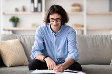 Gözlüklü genç bir adam kanepeye oturmuş, parlak aydınlatılmış oturma odasındaki kağıt yığınına notlar yazarken konsantre oluyor..