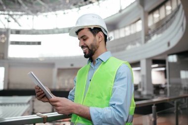 Yeşil yelek giymiş bir inşaat işçisi, inşaat halindeki bir binanın içinde tablet kullanırken gülümsüyor. İşiyle meşgul ve odaklanmış görünüyor..
