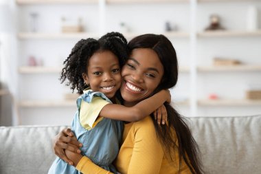Bir anne ve küçük kızı rahat bir kanepede kucaklaşırken mutlu bir şekilde gülümsüyorlar. Raflarla çevrili, aydınlık bir oturma odasında sıcak bir anın tadını çıkarıyorlar..