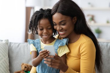 Bir anne ve küçük kızı parlak bir oturma odasındaki kanepeye yakın otururlar. Her ikisi de mutlu bir şekilde akıllı telefon ekranına bakar, gülümsemelerle dolu güzel bir anı paylaşır..
