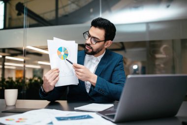 Takım elbiseli bir adam, çağdaş bir ofiste masa başında oturmuş renkli çizelgelere kağıt üzerinde işaret ediyor. Yakınlarda bir dizüstü bilgisayar ve belgeler var..