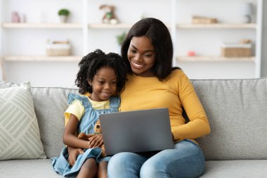 Genç bir anne ve kızı bir kanepede oturup dizüstü bilgisayardan bir şey izlerken gülümsüyorlar. Sıcak atmosfer, rahat bir ev ortamında birbirlerine olan yakınlıklarını gösteriyor..
