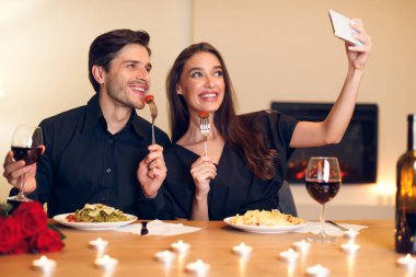 Evde romantik bir akşam, makarna, kırmızı şarap ve mum ışığının tadını çıkarırken gülümseyen ve selfie çeken bir çiftin samimi bir atmosfer yaratmasıyla geçer..