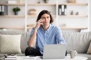 Önünde dizüstü bilgisayarı olan genç bir adam bir telefon görüşmesi yapıyor. Odaklanmış görünüyor ve rahat bir ev ortamı ile çevrili..