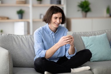 Genç bir adam kanepede bağdaş kurmuş oturuyor, akıllı telefonunu kullanırken gülümsüyor. Etrafında bitkiler olan parlak ve modern bir yaşam alanında rahat görünüyor..