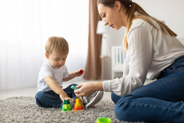 Genç bir çocuk ve bir anne renkli oyuncakları birlikte istiflemekten zevk alırken yumuşak bir halıya otururlar. Parlak odada doğal ışık var. Öğrenmek ve oynamak için sıcak bir atmosfer yaratıyor..