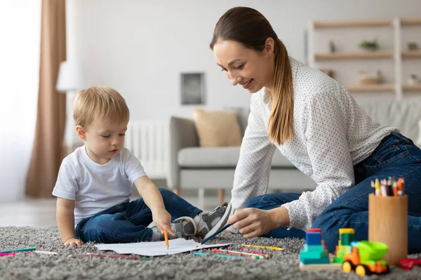 Mother and son are sitting on a soft carpet in a cozy living room. The boy is excitedly pointing at colorful drawings while the mother encourages him.
