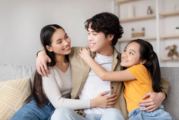 Genç bir adam kanepede iki gülümseyen kızla oturup sıcak anlarını paylaşıyor. Birbirlerini kucaklıyorlar, rahat ve davetkar bir yaşam alanıyla çevrili..