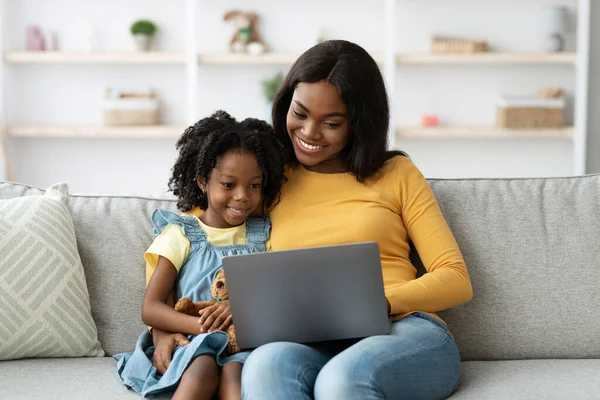 Genç bir anne ve kızı bir kanepede oturup dizüstü bilgisayardan bir şey izlerken gülümsüyorlar. Sıcak atmosfer, rahat bir ev ortamında birbirlerine olan yakınlıklarını gösteriyor..