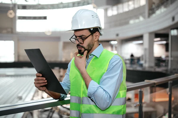 Güvenlik kaskı ve yansıtıcı yelek giyen bir adam açık alanları ve geniş pencereleri olan parlak, çağdaş bir ofis ortamında dururken bir belge üzerinde çalışıyor..