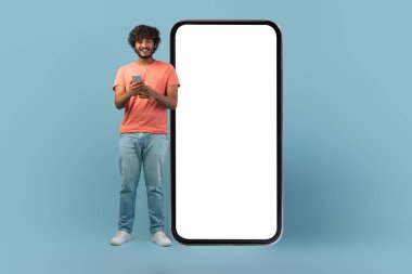 Neşeli genç bir Hintli adam boş ekranlı büyük bir cep telefonunun yanında duruyor. Akıllı telefonuyla meşgul, mavi bir stüdyoda gülümsüyor, canlı bir online teklif konsepti yaratıyor..