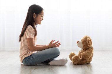 Genç bir kız, iyi ışıklandırılmış bir odada oyuncak ayısıyla mutlu bir şekilde bağdaş kurarak yerde oturuyor. Güneş ışığı perdelerin arasından akar ve sıcak bir atmosfer yaratır..