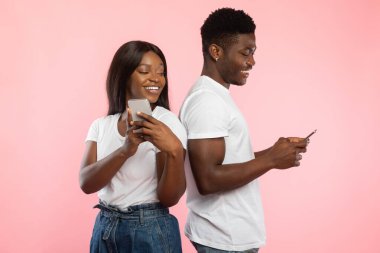 Cheerful black couple stands back to back, using their smartphones. They are in a playful mood, with a bright pink studio wall creating a lively atmosphere.