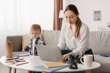 Bir anne laptopuna odaklanmış bir masada otururken küçük oğlu yakınlarda boya yapıyor. Konforlu oturma odasında doğal ışık ve neşeli bir dekor var. Sıcak bir atmosfer yaratıyor..