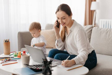 Bir anne dizüstü bilgisayarı üzerinde çalışırken gülümseyerek telefonda konuşur. Onun çocuğu yakınlardaki bir masada yapı taşlarıyla oynamaya odaklanıyor. Odanın parlak doğal ışığı var..