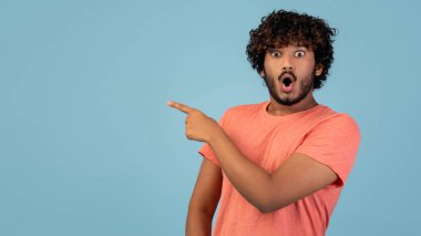 Kıvırcık saçlı, sıradan giyinmiş duygusal bir milenyum adamı mavi stüdyo arkaplanı üzerinde heyecanla fotokopi alanını işaret ediyor. Güçlü bir coşku taşıyor..