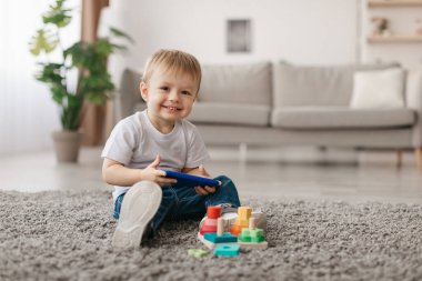 Neşeli genç bir çocuk sıcak bir oturma odasında lüks bir halıya oturmuş renkli blokları olan bir bina inşa etmeye odaklanmış. Parlak gülüşü oyun zamanındaki neşesini gösteriyor..