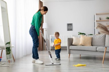 Bir anne, çocuğu yakınlarda durup merakla onu izlerken yerleri silmek için paspas kullanıyor. Güneş ışığı geniş oturma odasını doldurur..