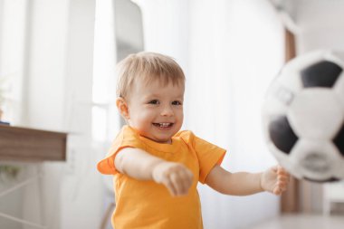 A cheerful young boy wearing an orange shirt is engaged in playful activity with a soccer ball indoors. The bright room has soft lighting, enhancing the joyful atmosphere.