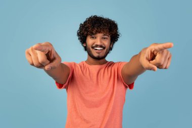 Kıvırcık saçlı ve sakallı neşeli bir genç adam gülümsüyor ve doğrudan kameraya bakıyor. Stüdyoda tek başına poz veriyor. Mavi bir arka planı var. Eğlenceli bir hava katıyor..