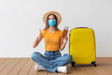 Yanında sarı bir bavulla yerde oturan bir kadın. Şapka ve maske takıyor, seyahat belgelerini tutuyor, başparmağını kaldırıyor, yolculuk için heyecanını gösteriyor..