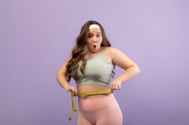 Şaşırmış bir Avrupalı kadın spor giyer ve belini bant ile ölçer. Sonuçtan dolayı çok heyecanlı ve stüdyo ortamında menekşe arkaplan karşısında duruyor..