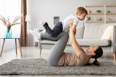 Bir anne yere yatar, gülümserken küçük çocuğunu yukarı kaldırır. Çocuk neşeli, rahat oturma odasında neşeli bir atmosfer yaratıyor..