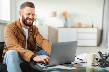 Genç bir adam rahat bir şekilde kanepede oturur, rahat bir oturma odasında dizüstü bilgisayarıyla çalışır. Modern ve parlak bir ortamla çevrili vazifelerine odaklanırken gülümsüyor..