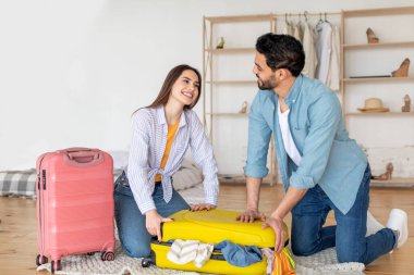 Birkaç kişi, rahat bir odada renkli bavullarını toplayarak yolculuklarına hazırlanıyorlar. Giysilerini düzenlerken ve yolculukları için heyecanlı bir şekilde gülümseme paylaşıyorlar..