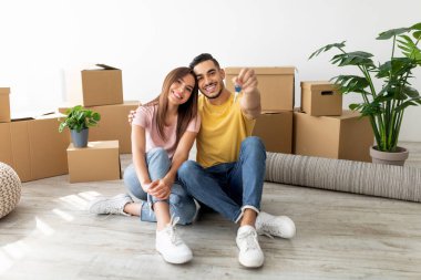 Mutlu çift yeni evlerinin zemininde oturup anahtarlarını gösteriyor. Etrafları taşınan kutularla ve kapalı alandaki bitkilerle çevrili, neşeli bir anı paylaşıyorlar..