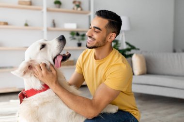 Parlak bir oturma odasında Golden Retriever 'ıyla neşeyle etkileşime geçen bir adam. Güçlü bağlarını ve mutluluklarını göstererek bir anlık şefkat gösterirler..