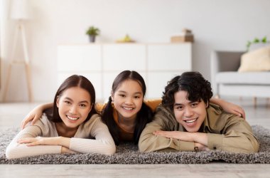 Üç aile üyesi, bir genç kız ve iki yetişkin, parlak bir oturma odasında yumuşak bir halının üzerinde yatıyor. Gülüyorlar ve eğleniyorlar, neşeli bir atmosfer yaratıyorlar..