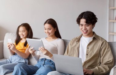 Three individuals are sitting comfortably on a couch, each focused on their devices. A girl is using a smartphone, while a woman and a man are using tablets and a laptop.