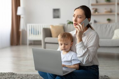 Bir anne telefonda konuşurken dizüstü bilgisayarla yerde oturuyor, endişeli görünüyor. Genç oğlu yanında, bir ekrana odaklanmış. Parlak, rahat bir oturma odasındalar..