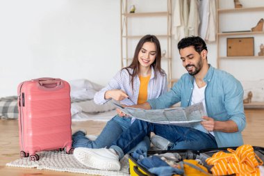 Genç bir çift, parlak ve rahat odalarının zemininde oturmuş haritayı incelerken gülümsüyor. Onlar seyahat planlarını tartışırken bavulları renkli giysilerle dolu..