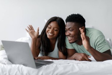 İki kişi bir laptopta yatakta yatarken keyifli bir video görüşmesi yapıyor. Gülümsüyorlar ve el sallıyorlar. Evlerinde sıcak, samimi bir atmosfer yaratıyorlar..