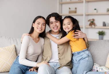 Bir erkek ve iki kız kardeş kanepeye yakın otururken mutlu bir şekilde gülümserler. Arka planda basit bir dekor ve sevgi dolu bir aile bağını simgeleyen sıcak bir atmosfer var..