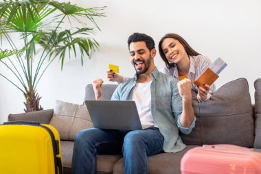 A man and a woman sit on a sofa, excitedly looking at a laptop as they book travel tickets. Luggage is nearby, showing their eagerness to travel.