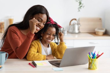 Bir anne ve kızı dizüstü bilgisayardan bir şey izlerken gülümsüyor ve kutlama yapıyorlar. Sıcak bir ev ortamında bir anlık neşeyi paylaşıyorlar..