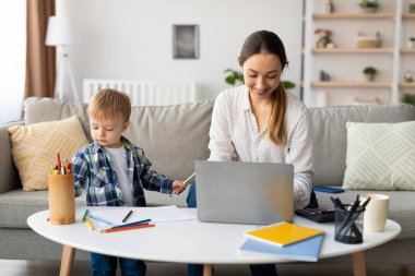 Bir anne, genç oğluyla evde iletişim kurar. Oğlan renkli kalemler ve kağıtlarla oynarken o laptopuna odaklanmış, sıcak ve çekici bir atmosferi gösteriyor..