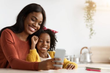 Bir anne ve küçük kızı, bir akıllı telefona bakarak bir anı paylaşırken gülümseyerek yakın otururlar. Kapalı mekanları parlak ve misafirperver, sıcak bir atmosfer yaratıyor..