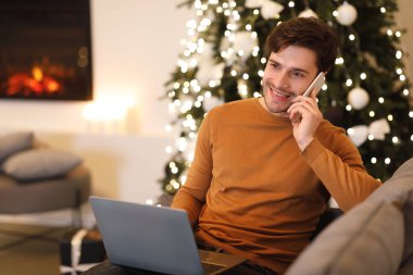 Kahverengi kazak giyen genç bir adam kanepede oturuyor, telefonuyla konuşuyor ve bir dizüstü bilgisayar üzerinde çalışıyor..